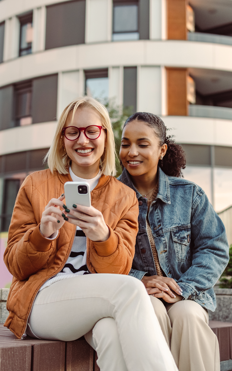 Zwei Frauen sitzen auf einer Stiege im freien, schauen auf das Mobiltelefon und lachen.