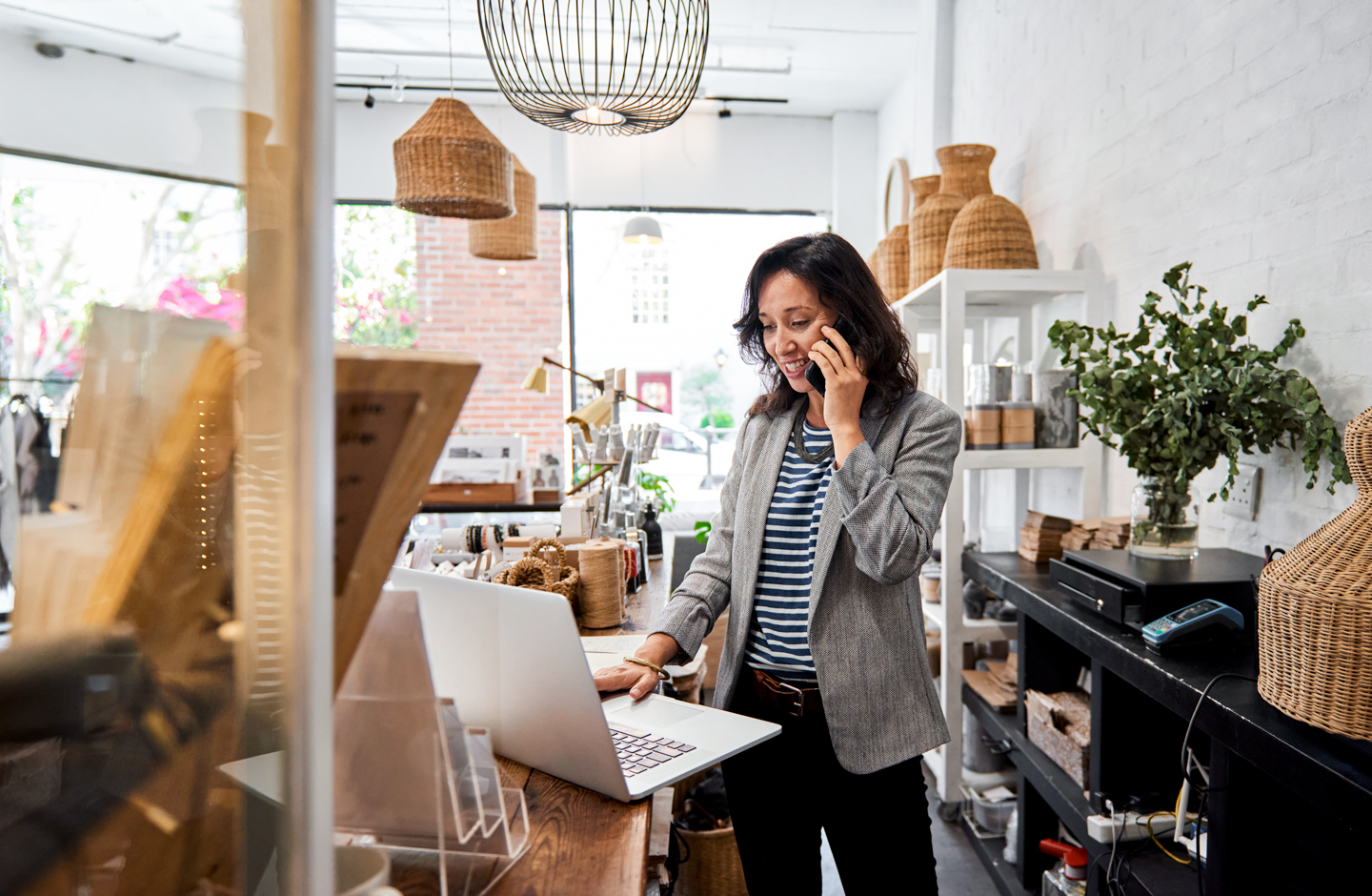 Eine Frau steht in ihrem Geschäft, telefoniert und tippt am Laptop